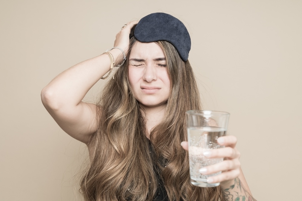 A young woman with a sleep mask drinking water with medicine after waking up with hangover